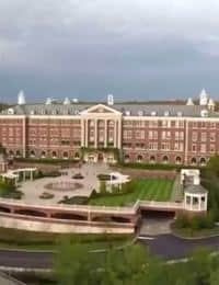 A large, stately brick building with white pillars and a grand entrance stands against a cloudy sky. It is flanked by well-manicured lawns, gardens, and a circular driveway. There are several smaller structures and greenery surrounding the main building.