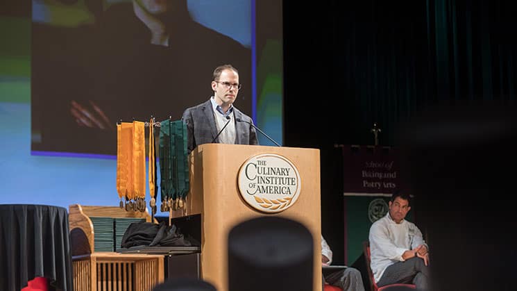 A person stands at a podium labeled "The Culinary Institute of America" giving a speech in an auditorium. Colorful banners hang in the background, and several seated individuals dressed in culinary attire are visible behind the speaker.