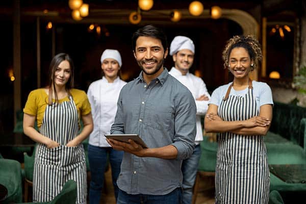 A group of five restaurant staff members are smiling at the camera. The man in front holds a tablet, while others wear aprons or chef hats, standing in a warmly lit dining area with green chairs and round hanging lights in the background.