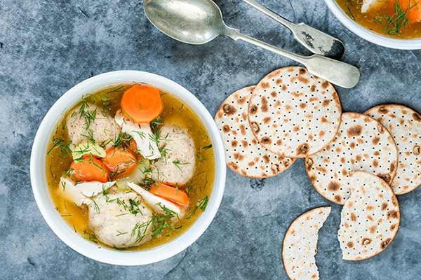 Top view of a bowl of matzo ball soup containing three matzo balls, chopped carrots, and herbs in a clear broth, placed on a grey surface. Next to the bowl are three round matzo crackers and two silver spoons.