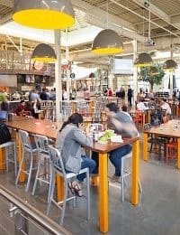 A bustling indoor cafeteria with high ceilings, showcasing long communal tables with bright yellow legs and gray metal chairs. People are seated, eating, and engaging in conversations. Large pendant lights hang from the ceiling, and a counter is visible in the background.