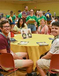 A group of people sit and stand around a round table covered with a yellow tablecloth. Some hold what appear to be magazines or brochures. They are in a brightly lit room with many others in the background. The atmosphere appears to be friendly and social.