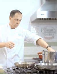 A chef in a white coat stands in a kitchen, focused intently as he cooks. He is using multiple pots and pans on a stovetop, with a vent hood and tiled backsplash visible in the background. His right arm is reaching towards a pot on the stove.