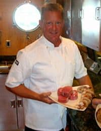 A chef in a white uniform is standing in a kitchen, holding a platter with various raw seafood, including fish and shrimp. There is a window in the background letting in natural light, and stainless steel cabinets are visible on the right.