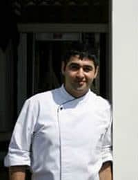 A chef dressed in a white chef's coat stands outdoors, partially leaning against a white wall. He has short, dark hair and a slight smile on his face.