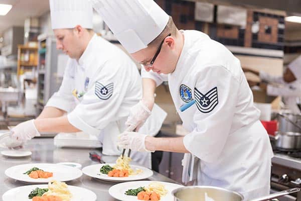 Two chefs in white uniforms and tall hats are plating dishes in a professional kitchen. One chef is garnishing a plate with precision, while the other chef is arranging ingredients. Plates of food with vegetables and pasta are on the counter.