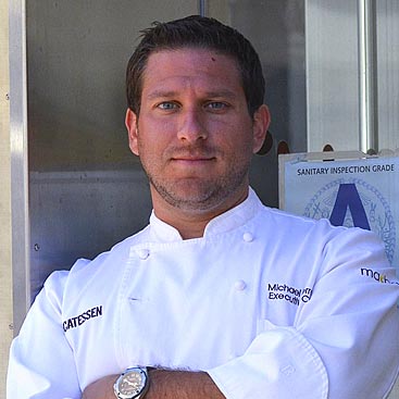 A man wearing a white chef's coat stands with his arms crossed in front of him. The coat has "Executive Chef" embroidered on it. He has short hair and is posing confidently. Behind him, a "Sanitary Inspection Grade A" sign is visible on the wall.