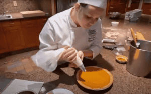 A chef in a white uniform and hat uses a piping bag to add cream or a similar topping to a pumpkin pie. The chef is focused on the task in a kitchen environment with wooden cabinets, a counter, and various baking tools and ingredients around.
