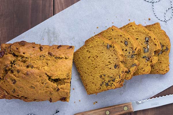 A sliced loaf of pumpkin bread with raisins or nuts is displayed on parchment paper on a wooden table. A serrated knife with a wooden handle is partially visible next to the bread.