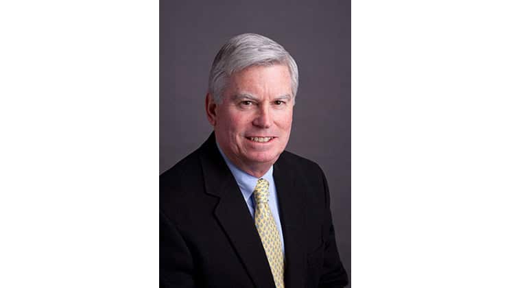 A middle-aged man with short white hair is dressed in a black suit, light blue shirt, and yellow patterned tie. He is smiling slightly and posing against a plain gray background.