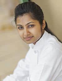 A young woman with dark hair pulled back, wearing a white chef's coat, gazes at the camera with a slight smile. The background is blurred, highlighting her focused and professional demeanor.