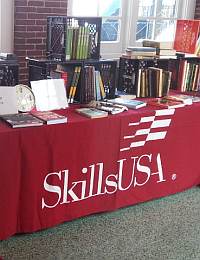 A table covered with a red tablecloth displaying the SkillsUSA logo is set up with books, folders, and various supplies. Crates in the background hold more books and binders. The setting appears to be indoors with windows in the background.
