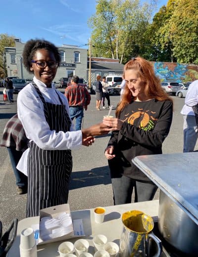 A person wearing a black and white striped apron and glasses is smiling and handing a small cup to another person with long reddish hair, dressed in a black shirt with a pumpkin graphic. They are outside, and other people and buildings are visible in the background.