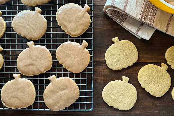 Pumpkin-shaped cookies cooling on a wire rack, with some placed on a wooden surface next to the rack. A checked cloth napkin and part of a yellow-lipped bowl are visible in the top right corner. The scene has a cozy, autumnal feel.