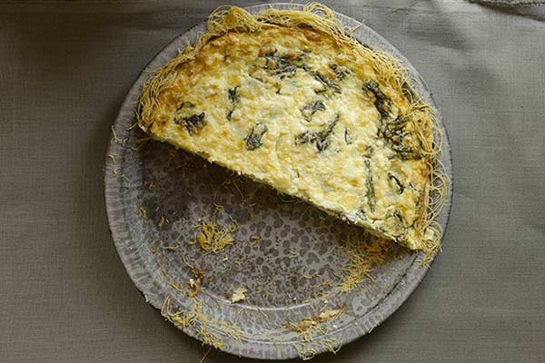 A round, partially eaten quiche with a crispy, golden crust and visible leafy green vegetables inside sits on a textured, gray plate. The quiche is missing a quarter slice, revealing its creamy interior with stringy, browned edges.