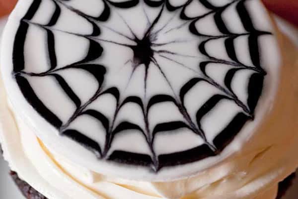 A close-up photo of a cupcake topped with white icing decorated with a spiderweb pattern in black. The design intricately extends from the center of the cupcake to the edges, creating a visually striking effect.