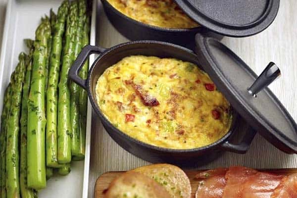 Two mini cast iron pots filled with baked vegetable frittatas, accompanied by a side of cooked asparagus, bread, and smoked salmon slices on a wooden surface. One of the pot lids is slightly ajar, and the contents appear fresh and appetizing.