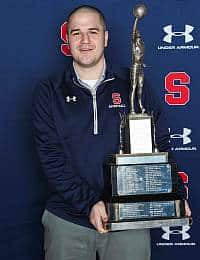 A person wearing a navy blue Under Armour jacket holds a large trophy with multiple tiers and a figure on top. They stand in front of a backdrop featuring a red "S" and Under Armour logos.
