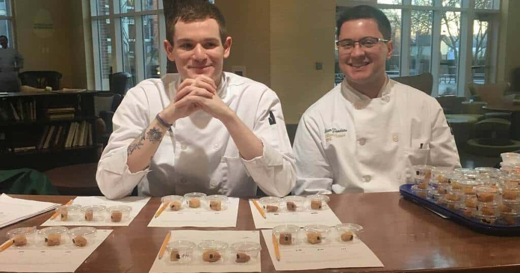 Two chefs in white uniforms sit at a table covered with small cups of food samples and papers. One chef smiles with his hands clasped, while the other grins, holding a pen. Both appear to be conducting a food tasting or evaluation.