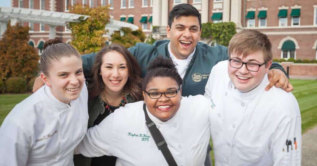 Five people are posing and smiling outside, with four of them dressed in white chef uniforms and one in casual clothing. They appear to be standing on a lush green lawn in front of a large brick building with white columns and green awnings.