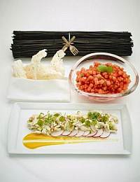 A culinary presentation features a rectangular plate with thinly sliced vegetables drizzled with a yellow sauce, a bowl of diced tomatoes with herbs, and a white napkin holding a piece of crispy bread. Black decorative sticks tied with a silver utensil set are in the background.