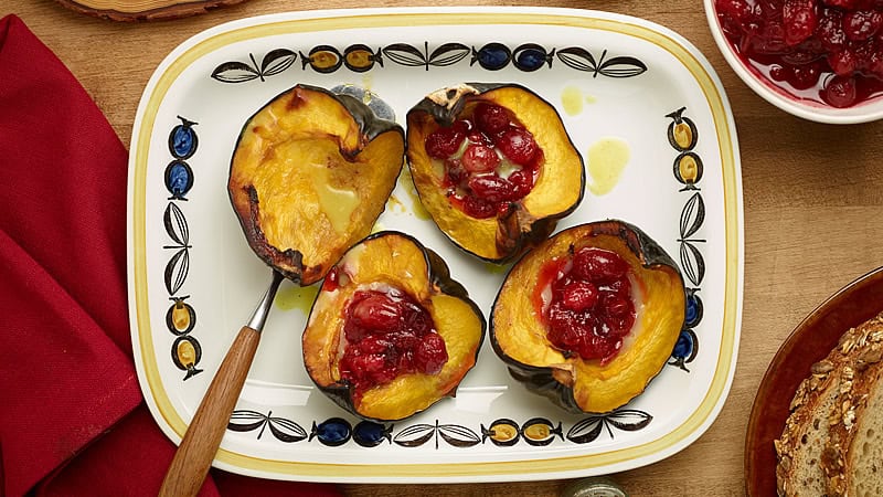 A rectangular platter holds four roasted acorn squash halves filled with a cherry and cranberry mixture. A red cloth napkin is partially visible on the left, and a small bowl of the cherry mixture is on the right. The platter is decorated with a leaf and fruit pattern.