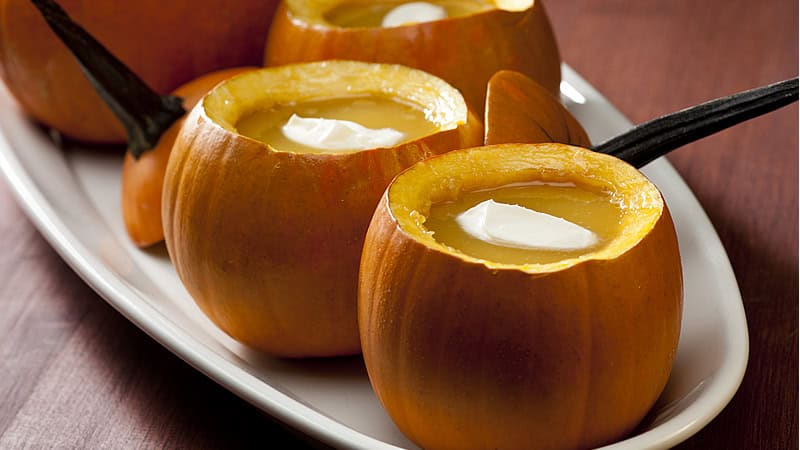 Small pumpkins filled with creamy soup, each topped with a dollop of sour cream, are arranged on a white serving platter. The pumpkins have their tops cut off and placed beside them, creating a festive and appetizing presentation.