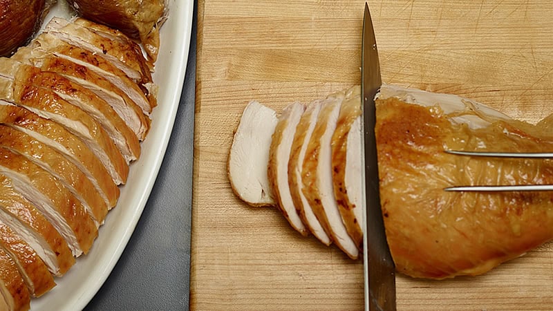 A close-up of a sliced, roasted turkey breast on a wooden cutting board. A knife is in the midst of slicing through the meat, held steady with a fork. A plate on the left side shows more slices of the turkey breast neatly arranged.