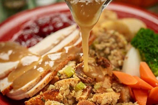A close-up of a plate of Thanksgiving food, featuring slices of turkey, stuffing, carrots, broccoli, potatoes, and cranberry sauce. Gravy is being poured over the turkey and stuffing.