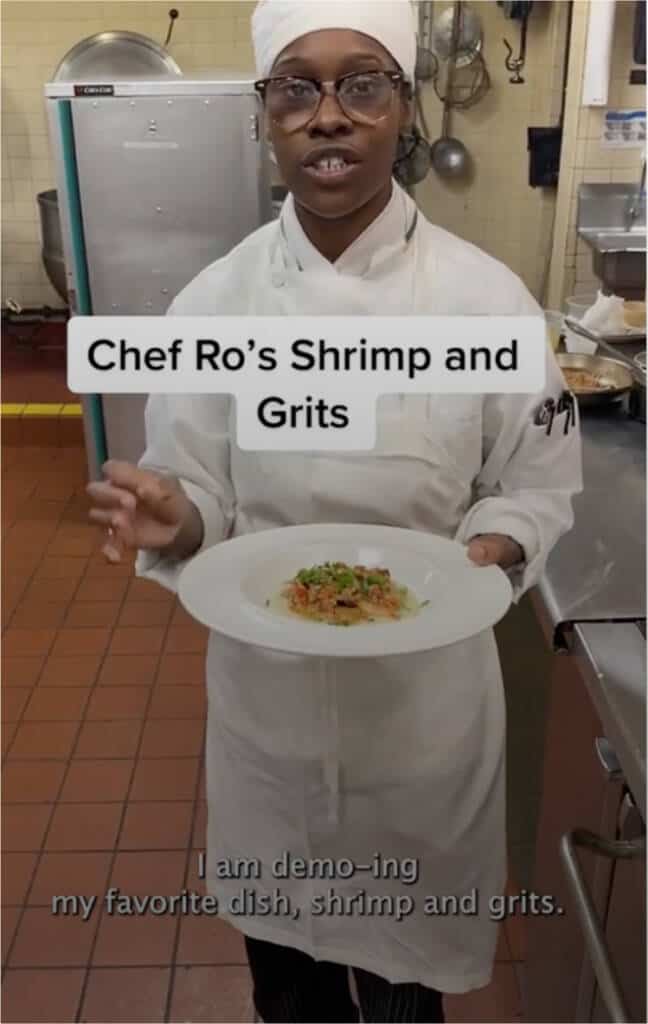 A chef in a white uniform and hat holds a plate of shrimp and grits in a kitchen. The text on the image reads, "Chef Ro’s Shrimp and Grits." The chef says, "I am demo-ing my favorite dish, shrimp and grits." Cookware is visible in the background.
