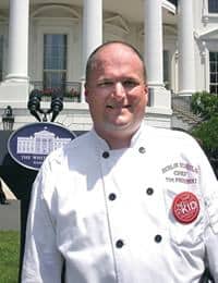 A person wearing a white chef's coat stands in front of a building that resembles the White House. The coat has embroidered text on the chest reading "Executive Chef" and features a red circular logo. A podium with the White House emblem is seen in the background.