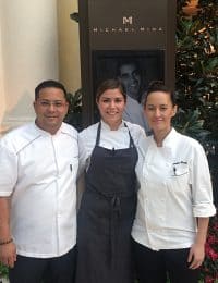 Three chefs stand in front of a sign with "Michael Mina" written at the top and a portrait of a man. They are wearing white chef coats, with the chef in the center also wearing a dark apron. They are posing closely together in a friendly manner.