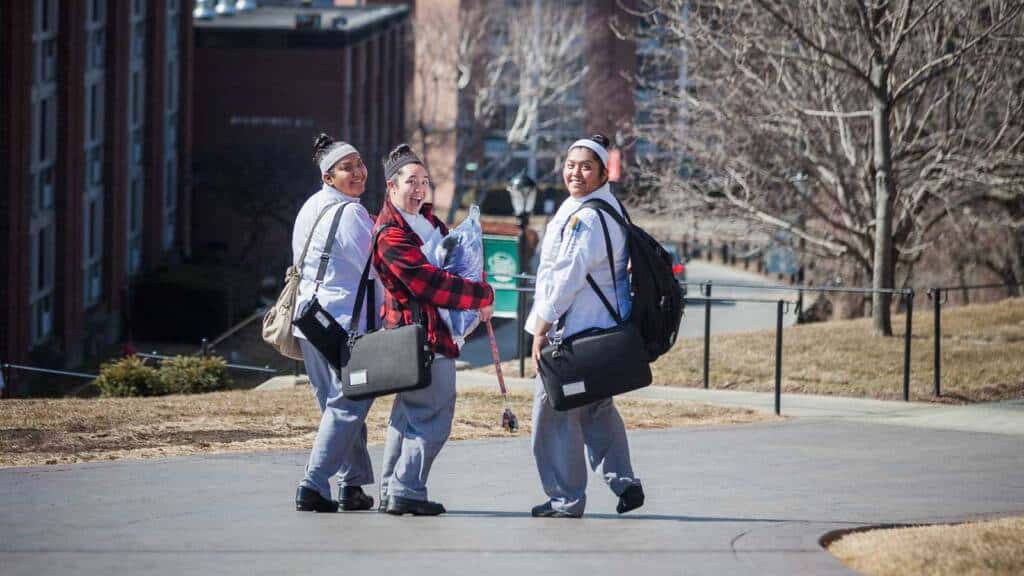 Three students in casual attire, carrying bags and looking back, walk down a paved path on a university campus. The background features brick buildings, trees, and a slightly overcast sky. The students appear to be cheerful and enjoying their walk.