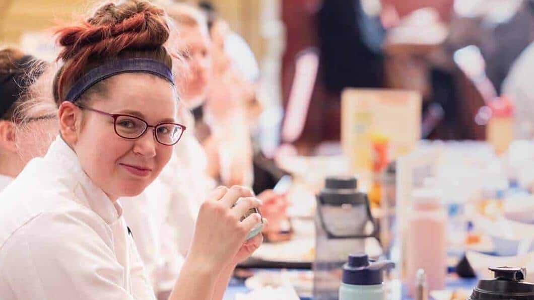 A person with glasses and a headband is seated at a table, smiling at the camera. They are wearing a white coat. Several other blurred figures are also seated at the table, and various bottles and containers are on the table in front of them.