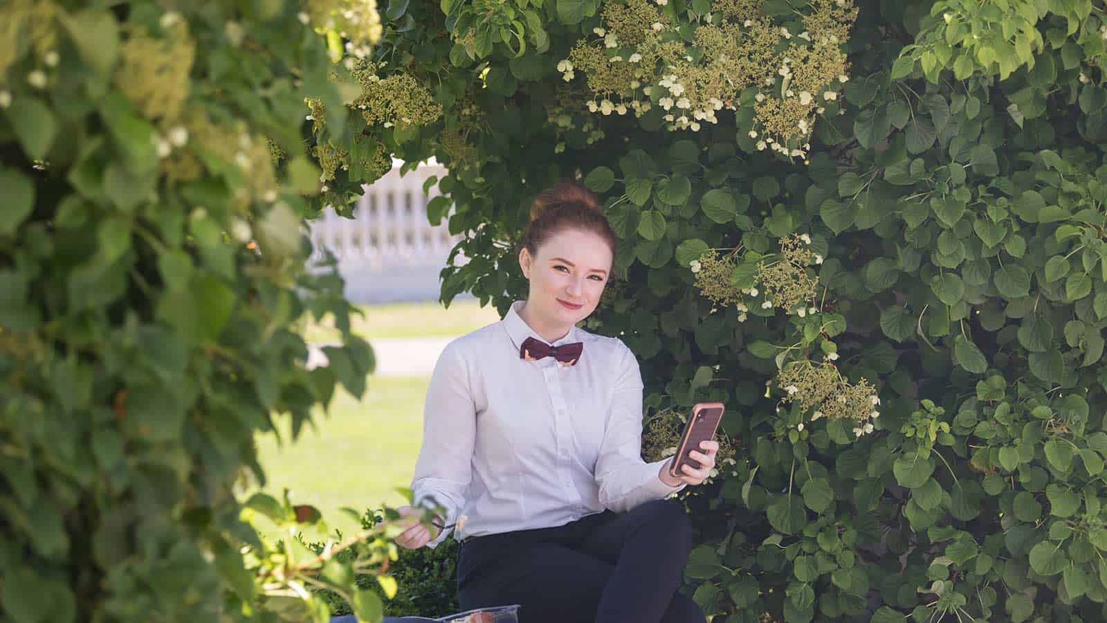 A person with a neat bun hairstyle is sitting outside surrounded by lush green bushes and white flowers. They are wearing a white shirt with a red bow tie and holding a smartphone in one hand while looking at the camera with a gentle smile.