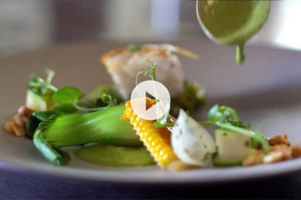 Close-up of an elegant plated dish featuring green vegetables, corn, nuts, a white sauce, and a piece of fish. Green sauce is being drizzled over the dish in the foreground. The plate is placed on a dark surface and there is a play button overlay in the center.