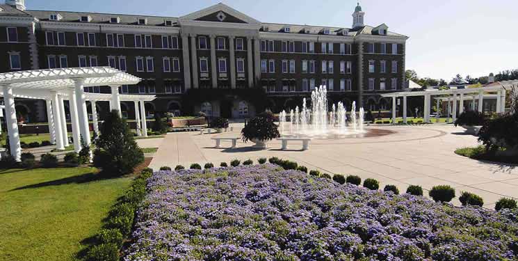 A large, historic building with a prominent central entrance stands behind a beautifully landscaped garden. The garden includes blooming purple flowers, paved walkways, white pergolas, and a grand fountain with water jets creating an elegant, inviting scene.