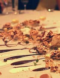 A close-up of a fancy dessert artfully plated on a white surface. The dish features crumbled elements, creamy dollops, colorful garnishes, and intricate drizzles of chocolate and white sauces. The background is out of focus, highlighting the dessert.