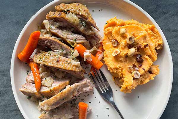 A plate with slices of roasted turkey, glazed carrots, and a serving of mashed sweet potatoes topped with nuts. A fork is placed next to the food on the plate.