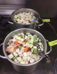 Two large metal pots filled with chopped vegetables and meat are simmering on a stovetop. The vegetables include cubes of green zucchini, white onion, and orange carrots. Both pots have ladles resting on the handles, with labels attached.