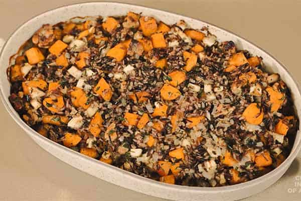 A white, oval baking dish filled with a mixed vegetable casserole. The dish prominently features diced sweet potatoes, wild rice, diced onions, and other mixed vegetables. The top is slightly browned and crisp from baking.