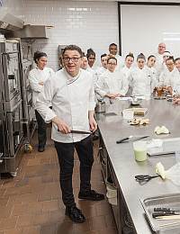 A chef stands at the head of a stainless steel counter in a commercial kitchen, holding a knife. A group of culinary students in white uniforms gather around him, attentively watching and listening. Kitchen equipment and ingredients are visible in the background.