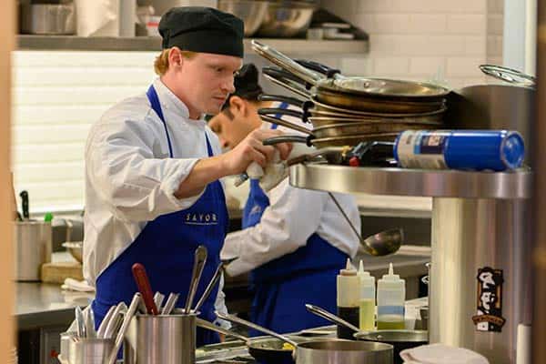 A chef wearing a black hat and a blue apron is preparing food in a professional kitchen. He is focused on his task, with various cooking utensils and bottles of sauces around him. Another chef is working in the background.