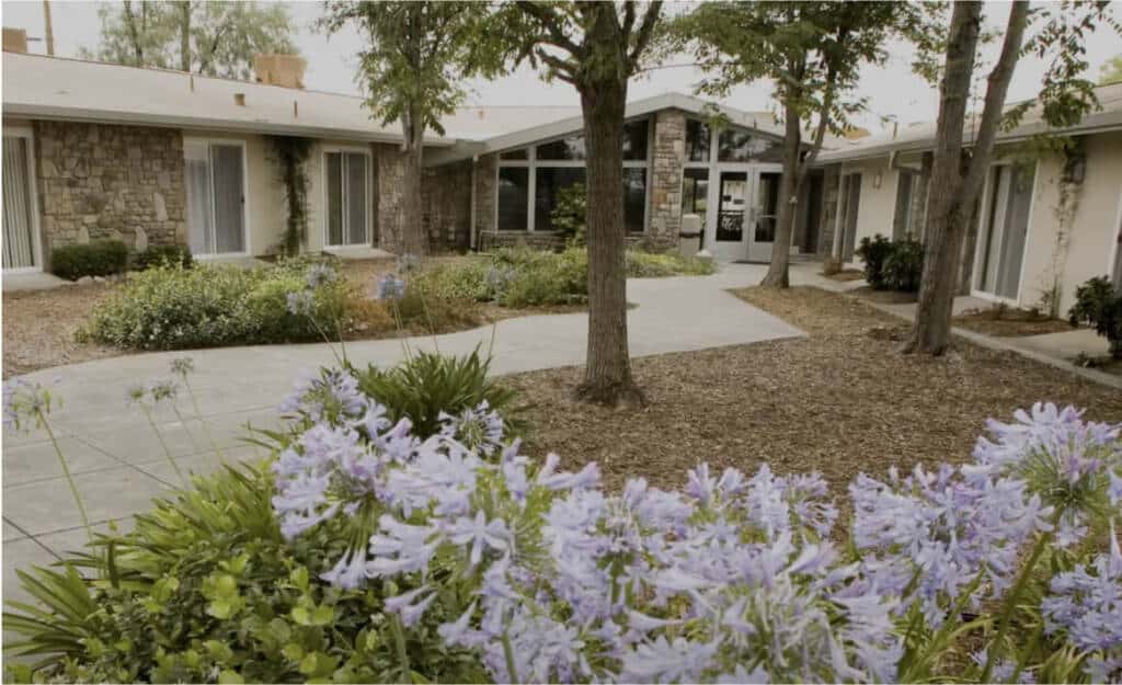 A courtyard of a single-story building features a central walkway surrounded by trees and flower beds with blooming purple flowers. The structure showcases stone and wood detailing, large windows, and a mix of paved and landscaped areas.