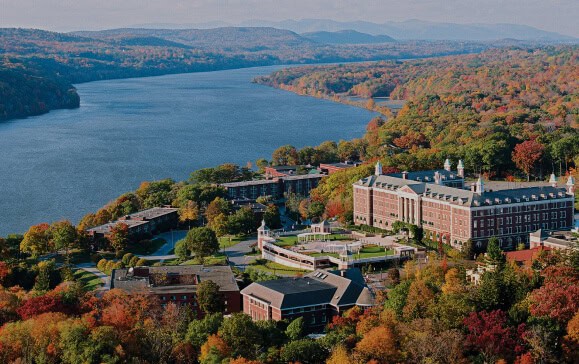 Aerial view of a large campus with multiple buildings surrounded by trees in autumn colors. The campus is bordered by a wide river on one side, with rolling hills visible in the distance under a clear sky.