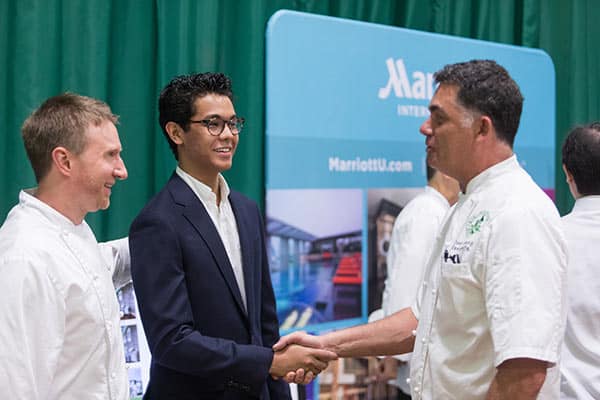 A woman in a black blazer shakes hands with another woman wearing a white chef coat at a career fair, reflecting the vibrant campus life at The Culinary Institute of America. Both are smiling, while another person in a white chef coat stands nearby. Other attendees and booths are visible in the background.