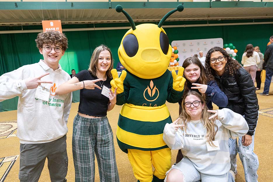 Image of smiling CIA students standing with Sting, CIA’s honeybee mascot in the gymnasium.
