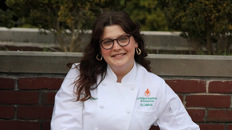 Image of CIA student Annie Milostan in her graduation jacket sitting in front of a brick wall.