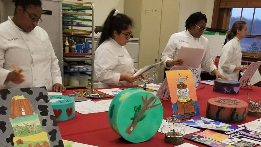 Three chefs in white coats are standing behind a table displaying colorful and artistic culinary creations. The table is covered with a red cloth, and various decorated culinary items, including cakes and edible art, are arranged on it. Shelves in the background have various books and supplies.