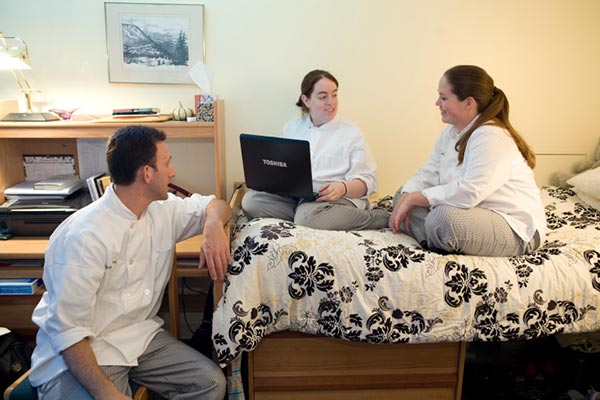 Image of three CIA students hanging out in a dorm room on campus. Two women are sitting on the bed while a man is sitting in a chair at the desk.